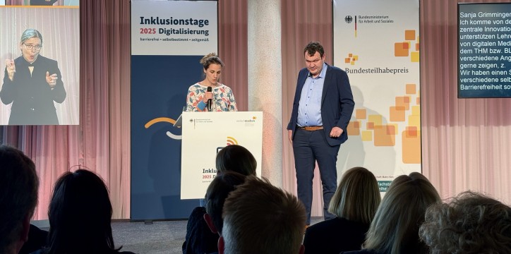 Sanja Grimminger und Andreas Deitmer stehen mit Mikrophon auf der Bühne. Im Hintergrund sieht man auf Bildschirmen schriftsprachliche sowie Gebärdendolmetschung.