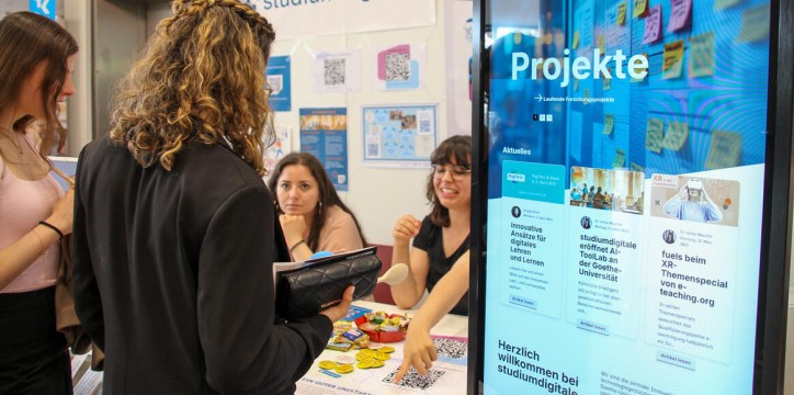 Eine Person mit lockigem Haar, die einen schwarzen Blazer trägt, steht an einem Informationsstand mit dem Titel